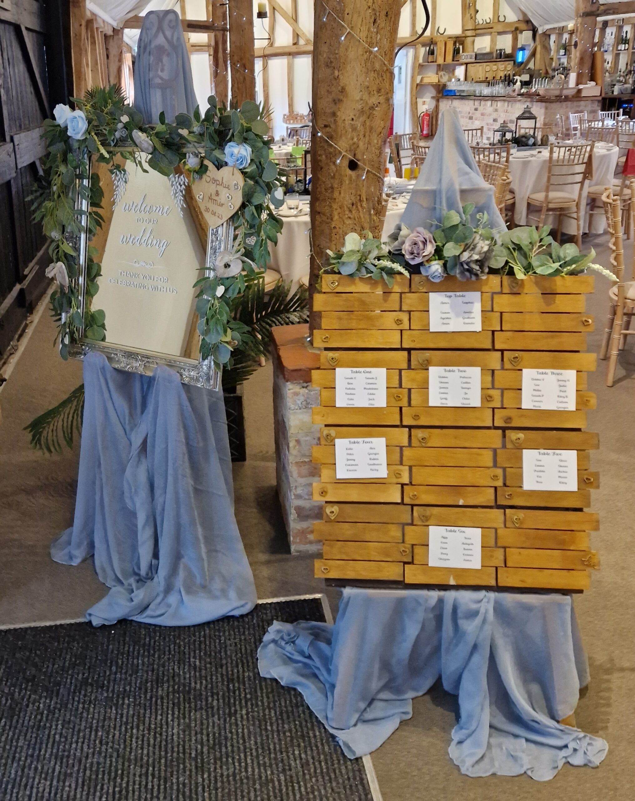 Silver mirror welcome sign and wooden board table plan, both with dusky blue swags and flowers